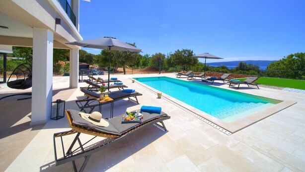 A modern outdoor pool area with lounge chairs and umbrellas, surrounded by a landscaped garden and a view of the sea. - Villa MAGNIFICA with pool (Pinezići)