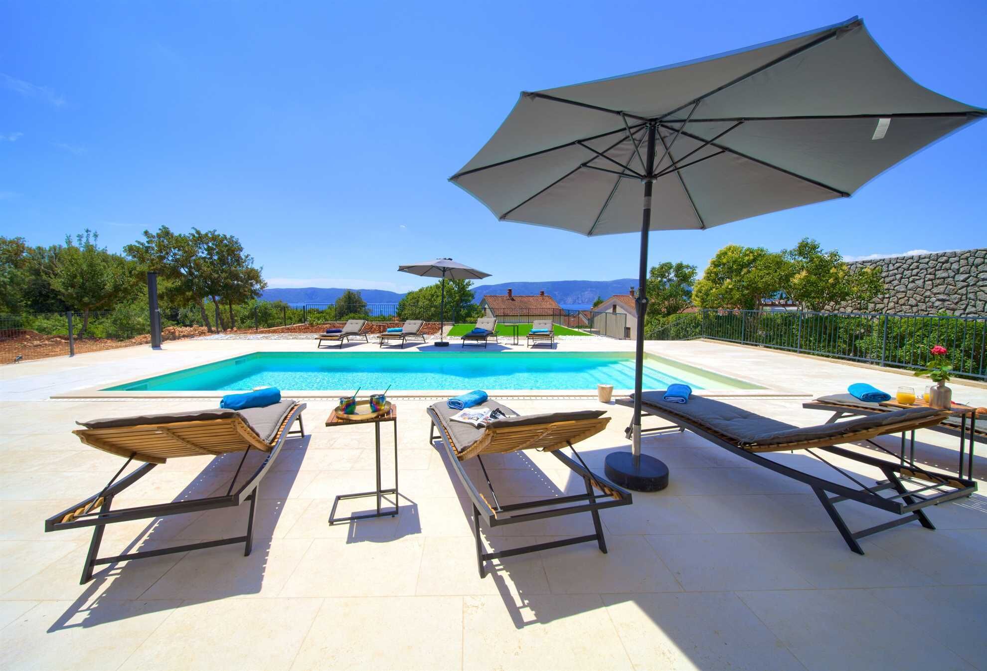 Un espace aménagé au bord de la piscine, avec des chaises longues et des parasols, une piscine à l’eau d’un bleu limpide et une vue sur les montagnes en arrière-plan.