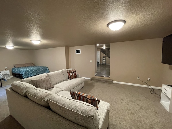 A spacious living room with a large beige sectional sofa, a blue bed with patterned bedding, and a staircase leading to an upper level.