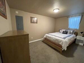 A bedroom with a bed, a nightstand, a dresser, a door, a framed picture on the wall, and a window with curtains.