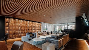 A modern interior with a wooden ceiling, a seating area with sofas and ottomans, a wooden wall with glass panels, and a large window providing natural light. - Nido x Neuchatel Polanco (Mexico City)