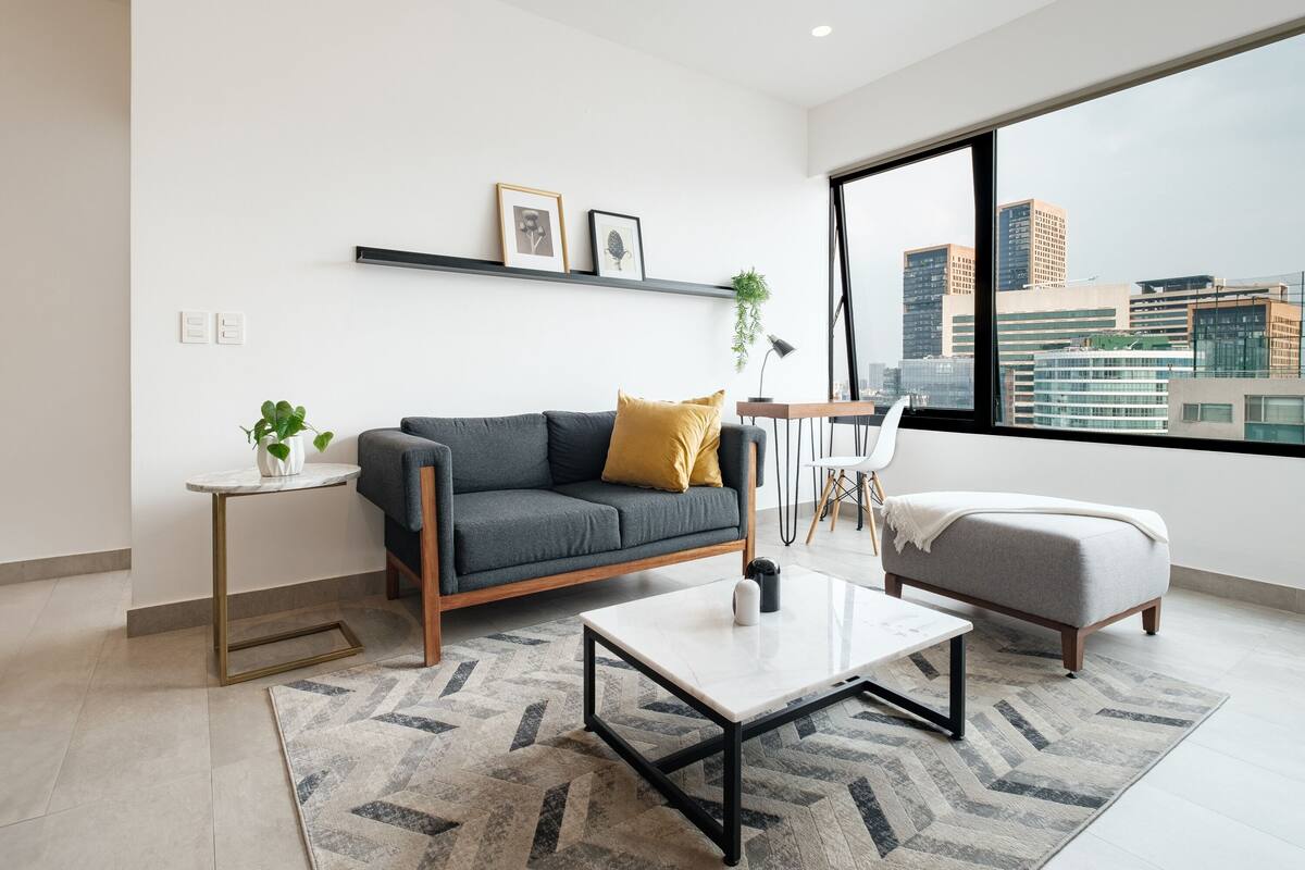Ein modernes Wohnzimmer mit einem grauen Sofa, einem weißen Couchtisch und einem grauen Hocker. Durch ein großes Fenster ist die Stadt zu sehen, ein kleiner Beistelltisch mit einer Pflanze und ein Regal mit gerahmten Bildern.