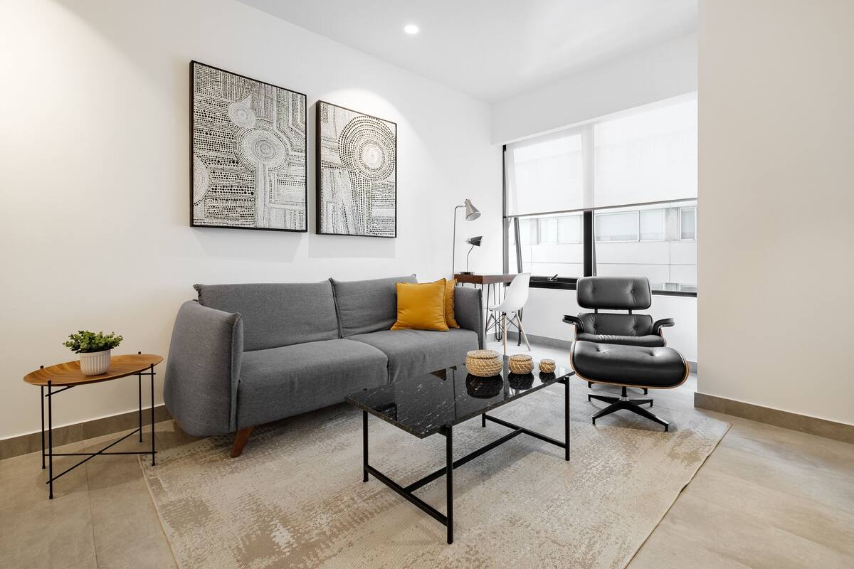 Ein modernes Wohnzimmer mit einem grauen Sofa, einem schwarzen Couchtisch, einem schwarzen Bürostuhl und zwei abstrakten Wandbildern.