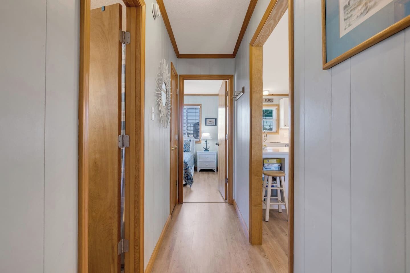 Un couloir étroit, avec des portes en bois, donnant sur une chambre où l’on aperçoit un lit, une table de chevet et une lampe.