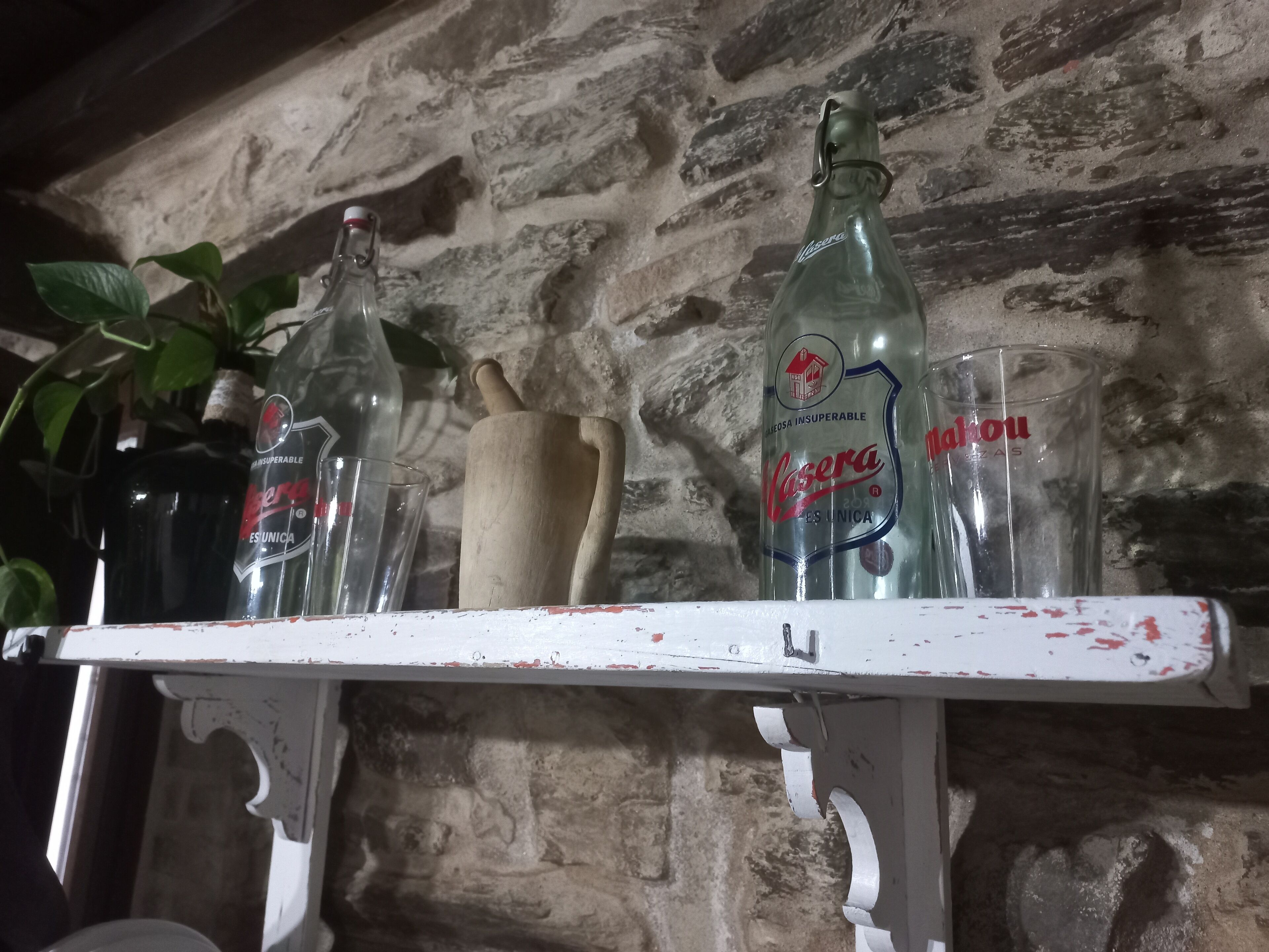Une étagère avec une bouteille de « Naser » et un rouleau à pâtisserie en bois, le tout adossé à un mur de pierre.