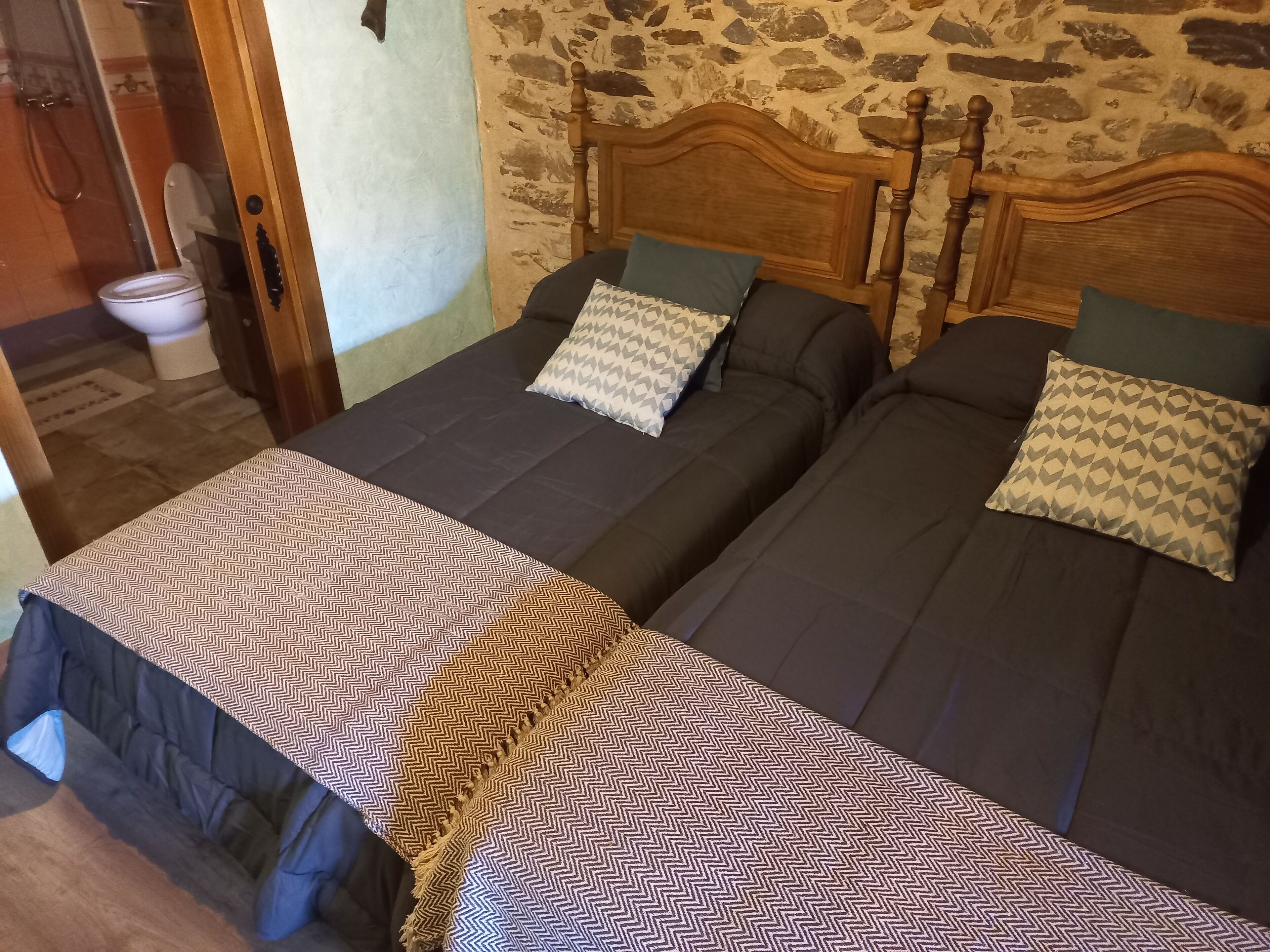 A room with two single beds, a wooden headboard, a patterned bedspread, and a stone wall.