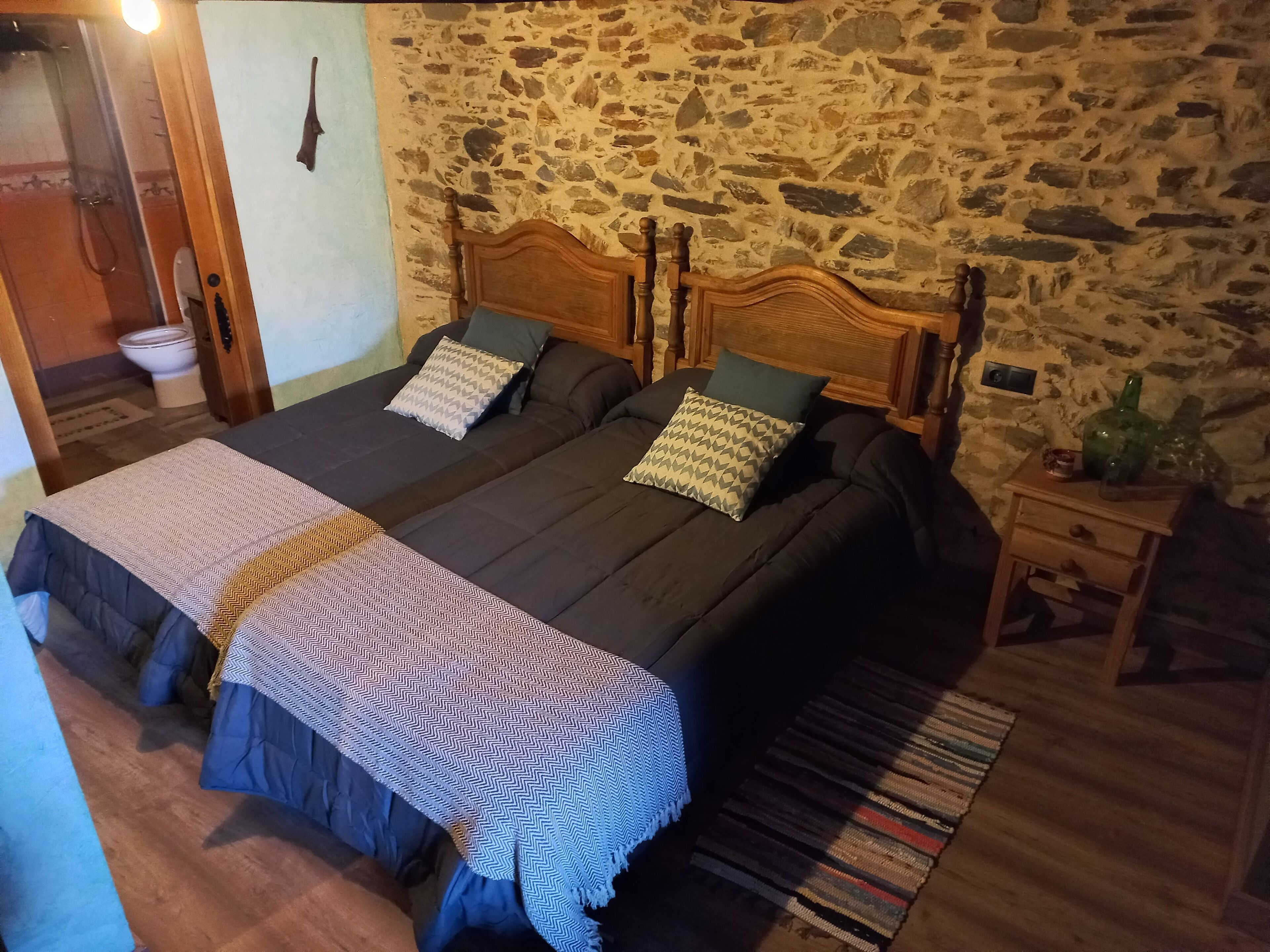 Una cama doble con cabecera de madera, dos almohadas y una colcha estampada. Una mesita de noche de madera con un jarrón y una botella están junto a la cama. Se ve una pared de piedra y una puerta de madera al fondo.