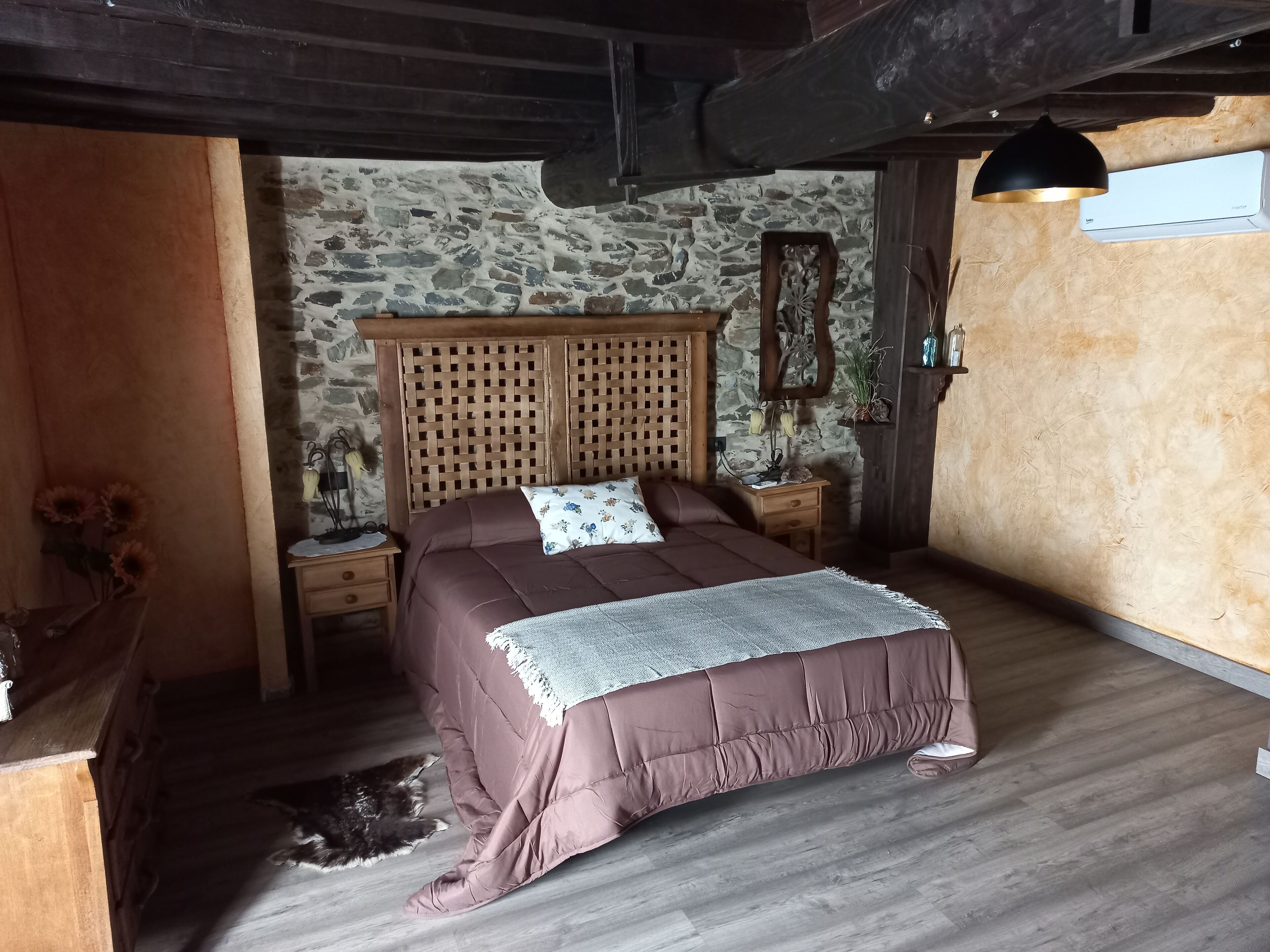 A bedroom with a wooden headboard, a stone wall, a wooden floor, a bedside table with a lamp, and a cat on the floor.