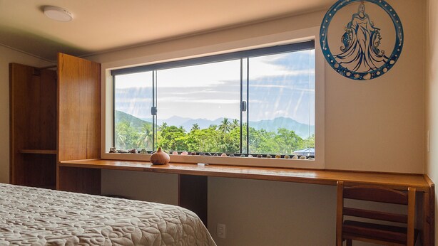 Une chambre avec une grande fenêtre, un lit recouvert d’une couvre-lit à motifs, une table de chevet en bois et une assiette décorative accrochée au mur.