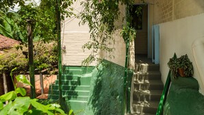 Un escalier avec des rampes vertes menant à une porte, le tout entouré de verdure et d’un mur de briques.