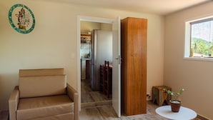 Une pièce avec un canapé beige, un meuble en bois, une petite table sur laquelle est posée une plante en pot, et une vue sur une cuisine par une porte ouverte.