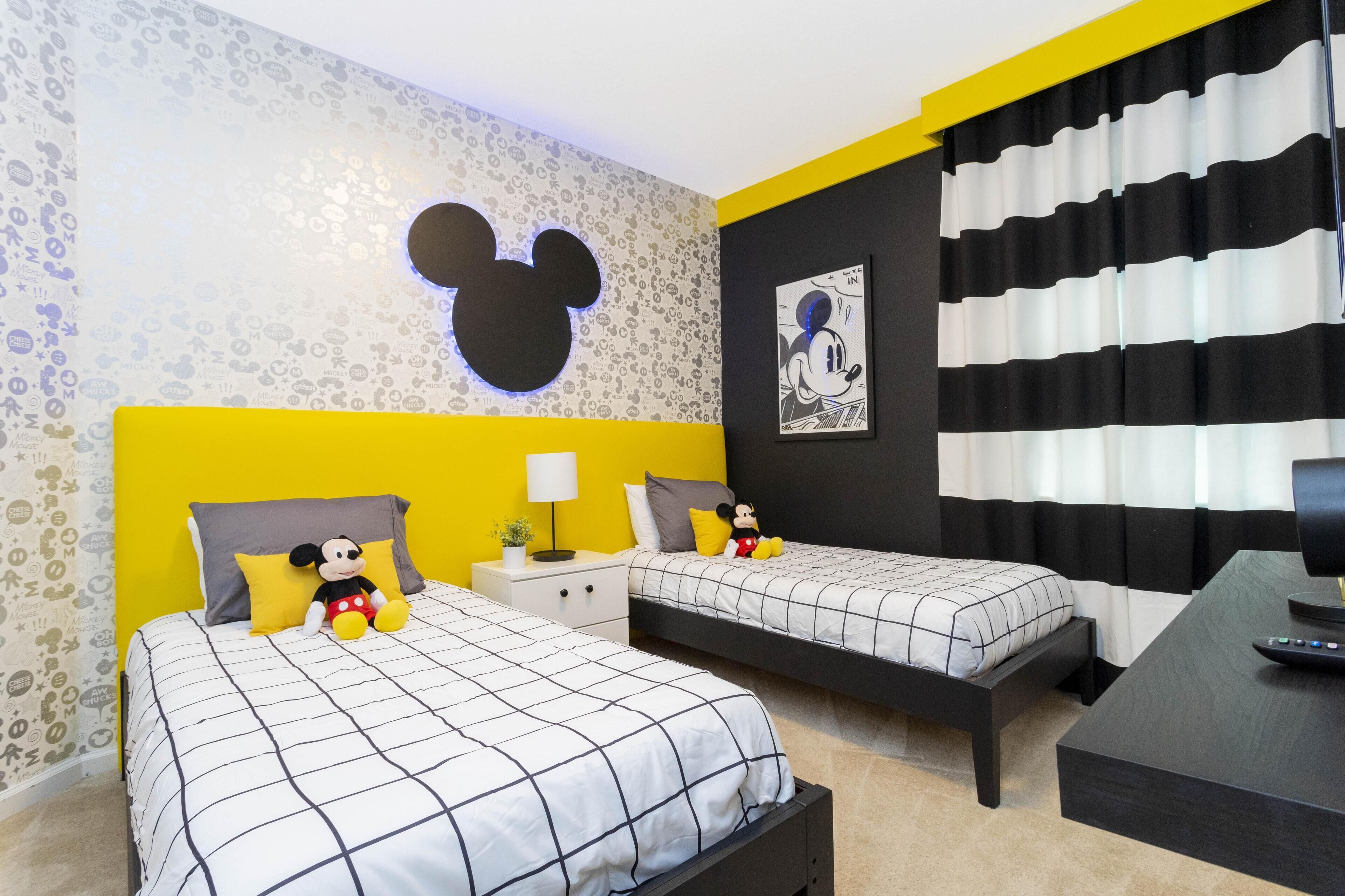 Habitación de hotel con dos camas, cabecera con la figura de Mickey Mouse, un peluche de Mickey y Minnie, cortina a rayas negras y blancas y un control remoto sobre una mesa.