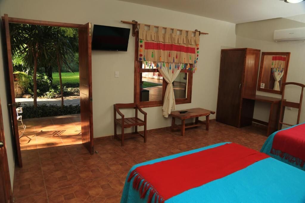 Una habitación con cama, escritorio, silla y televisión. Se ve una ventana con cortinas y vistas a la vegetación.