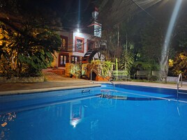 A swimming pool with a metal ladder, surrounded by plants and a building with a red and white facade.
