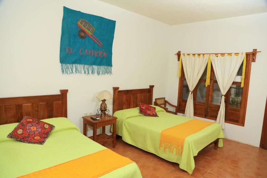 A room with two beds, a wooden headboard, a nightstand with a lamp, a window with curtains, and a blue banner on the wall.