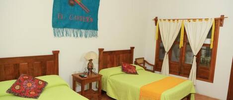 Una habitación con dos camas, cabecero de madera, mesita de noche con lámpara, ventana con cortinas y un cartel azul en la pared.