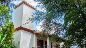 Edifício de dois andares com varanda, rodeado por vegetação exuberante e céu claro.