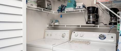 Un lavadero con dos lavadoras y secadoras de carga frontal blancas, estantes con toallas dobladas, una cafetera y una caja de detergente Tide.