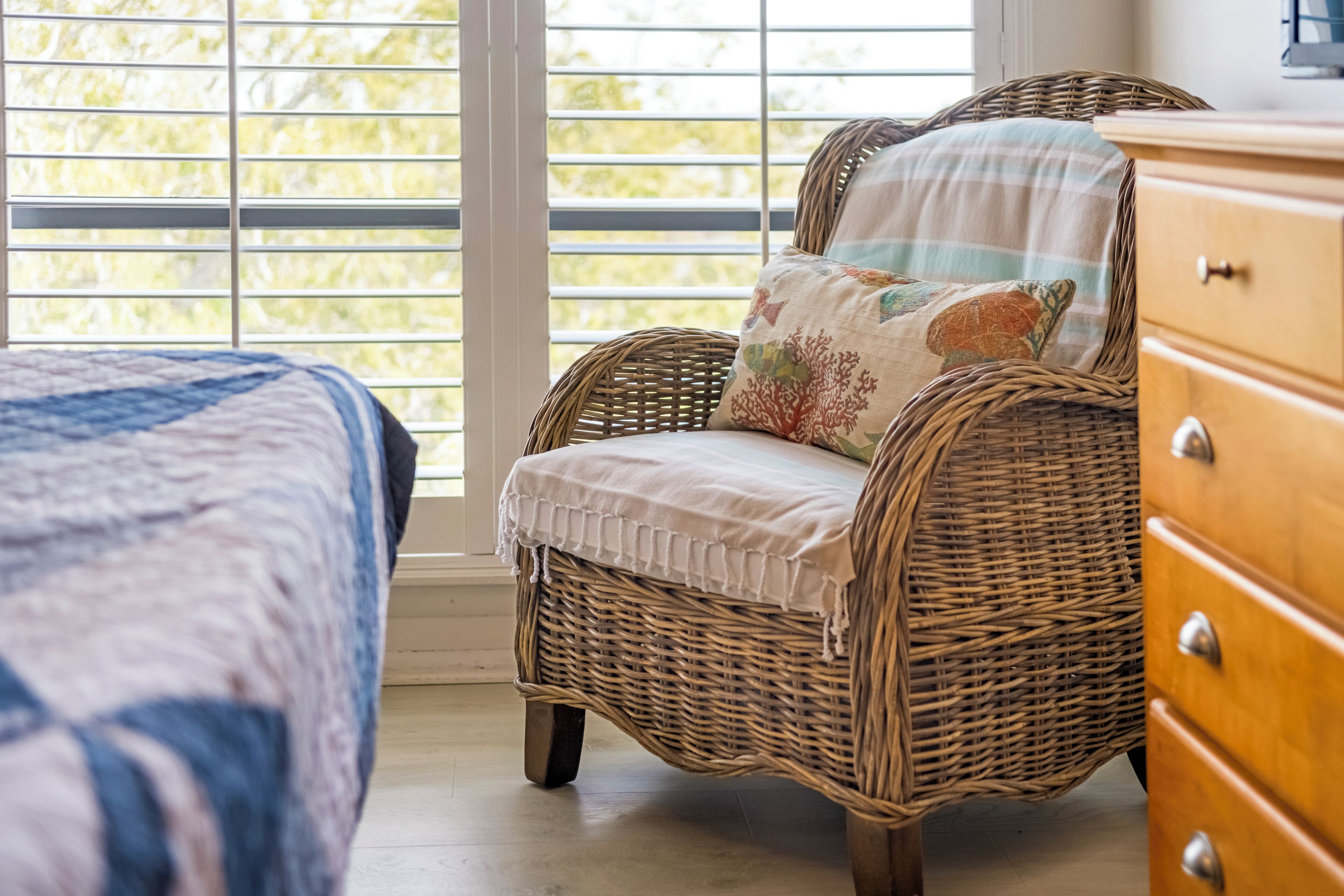 Une chaise en osier avec un coussin et une commode en bois, dans une pièce dotée d’une grande fenêtre.