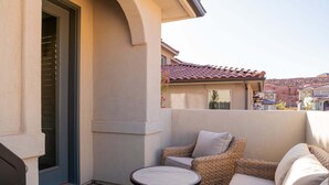 Eine Terrasse mit rundem Tisch und Korbmöbeln, eine Tür mit Fenster und ein Gebäude mit Ziegeldach im Hintergrund.