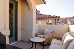 Un patio con una mesa redonda y muebles de mimbre, una puerta con ventanal y un edificio con tejado de tejas al fondo.