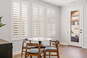 Un comedor con una mesa redonda y cuatro sillas de madera, una puerta blanca que da a una vista de un edificio, y una ventana con contraventanas blancas.