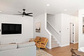 A modern living room with a flat-screen TV, a ceiling fan, a staircase, and a door leading to another room.