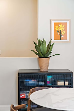 Quarto com planta em vaso sobre um móvel, quadro pendurado na parede e mesa com cadeira.