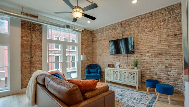 A living room with a brick wall, a leather sofa, a blue armchair, a white cabinet, and a TV mounted on the wall. - Majestic Lofts #201- Printers Alley Area (Nashville)