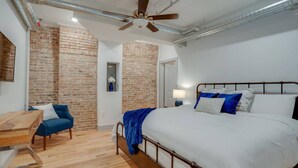 A bedroom with a large bed, a wooden desk, a blue chair, and exposed brick walls. - Majestic Lofts #201- Printers Alley Area (Nashville)