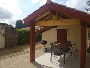 A covered patio area with a wooden table and chairs, a barbecue grill, and a wooden door. - Renovated Landes house in the heart of the village (Lit-et-Mixe)