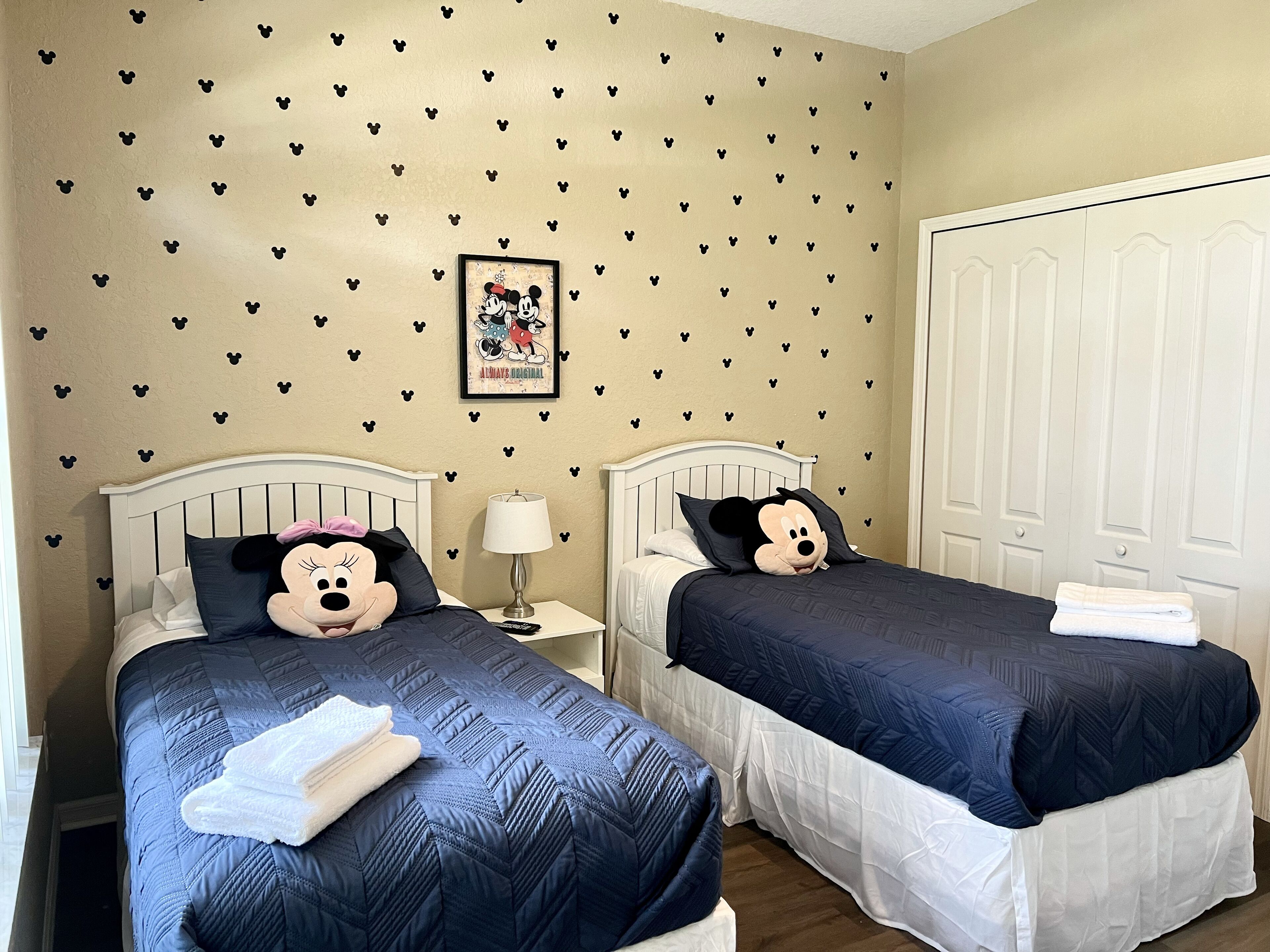A hotel room with two beds, each with a Mickey Mouse pillow, a framed Mickey and Minnie Mouse poster, and a white door.
