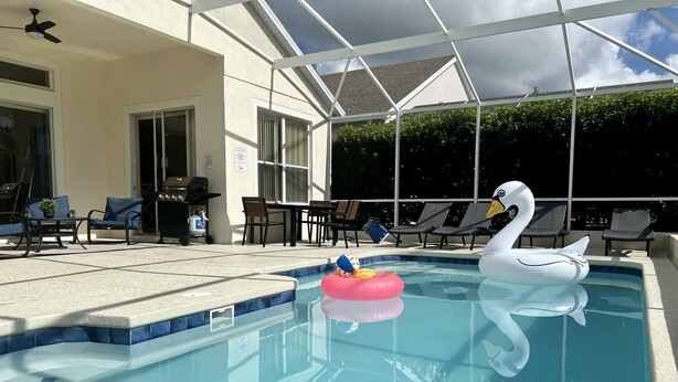 En swimmingpool med en svanebåd og en lyserød bold, omgivet af liggestole og en patio med grill.
