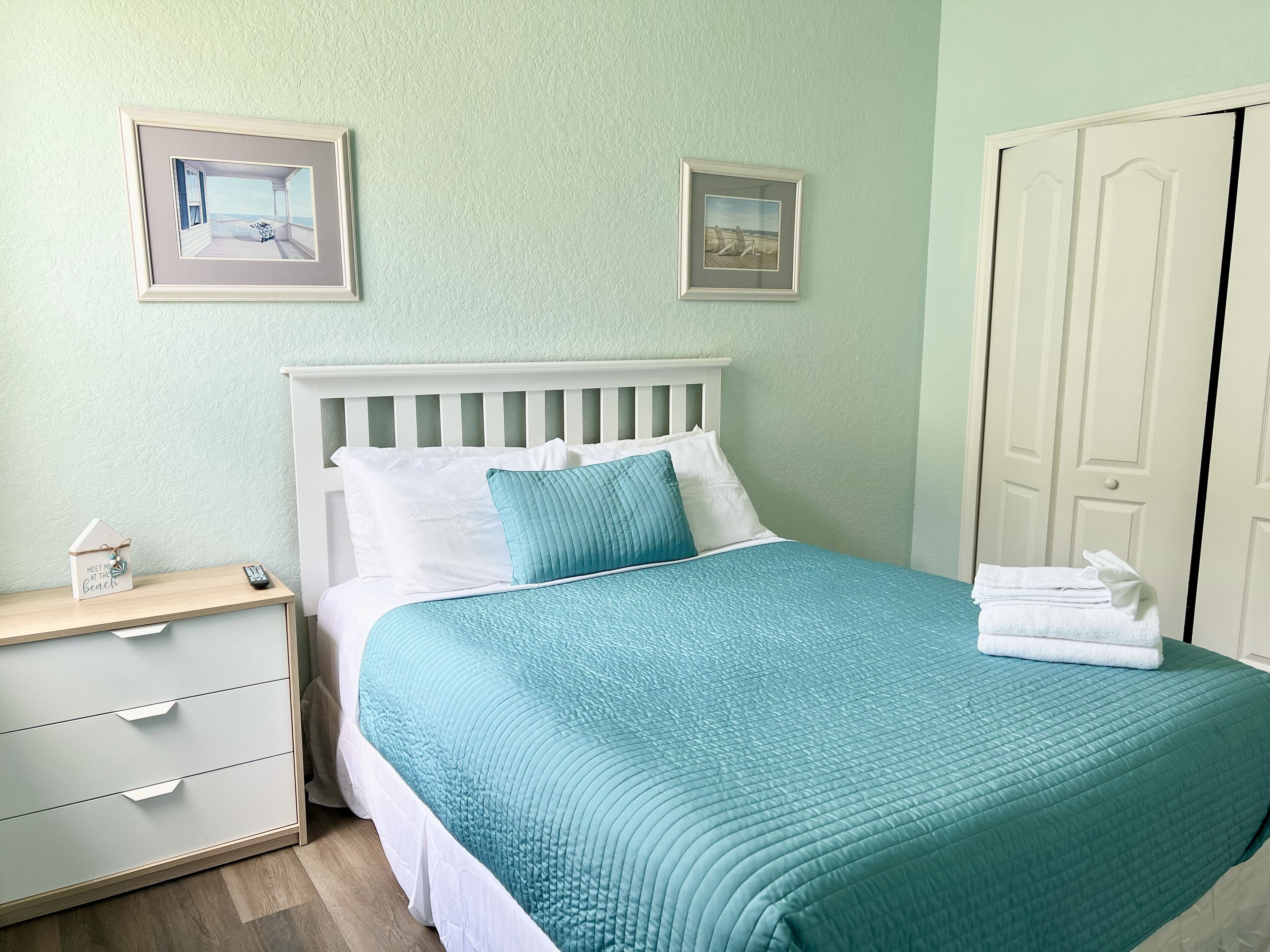 A neatly made bed with a teal bedspread, a white headboard, and two framed pictures on the wall.