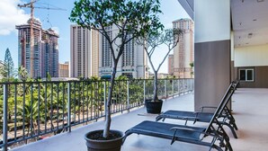 Un balcon agrémenté de deux fauteuils et de plantes en pot, d’où l’on peut admirer un paysage urbain avec de hauts immeubles et des grues de construction.