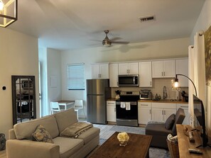 A living room with a sofa, coffee table, and a TV. There is a kitchen area with appliances and cabinets.