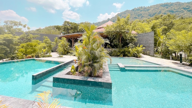 Un área de piscina con agua azul cristalina, rodeada de exuberante vegetación y montañas al fondo.