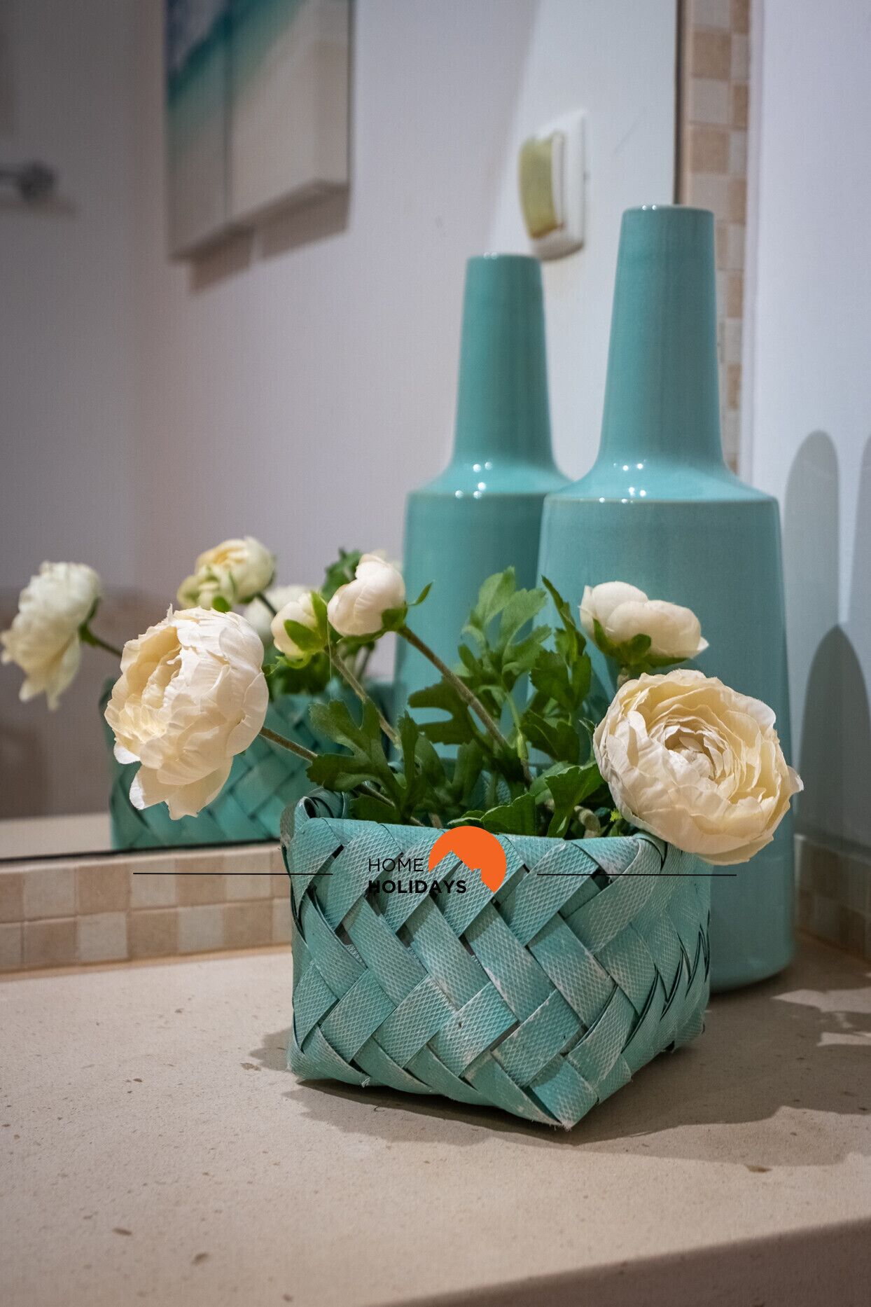 Ein Badezimmer mit einem Spiegel, zwei türkisfarbenen Vasen und einem gewebten Korb mit weißen Blumen.