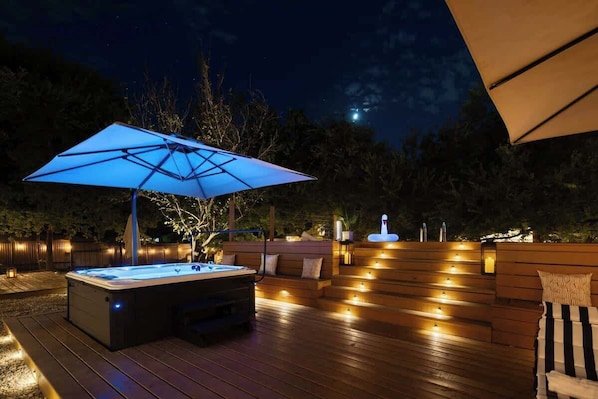 Un jacuzzi con una sombrilla azul, terraza de madera y zona de estar al aire libre por la noche.