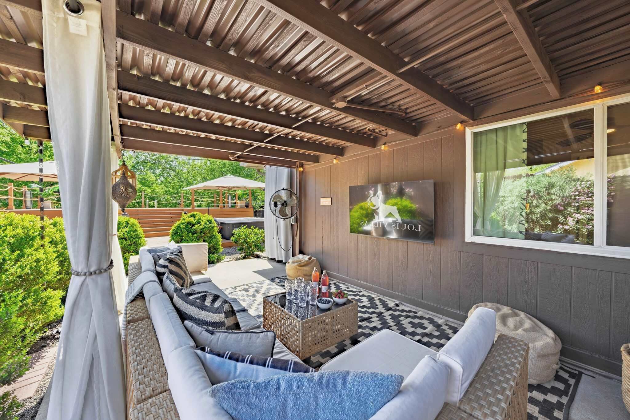 A covered outdoor seating area with a sofa, cushions, a woven coffee table, and a wall-mounted TV.