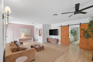 Una sala de estar moderna con sofá, mesa de centro y televisor. Se observa una puerta corrediza tipo granero y un ventilador de techo.