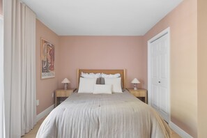 A bedroom with a bed, two bedside tables, a nightstand, a door, and a wall with a framed picture.