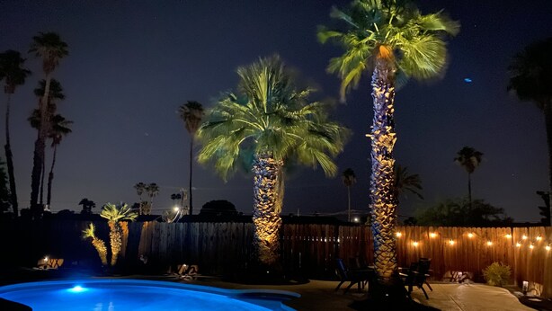 Un espacio al aire libre iluminado con luces colgantes, piscina, y palmeras.