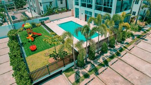 Un edificio moderno con piscina, rodeado de palmeras y un área de juegos cercada.