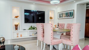 Un comedor moderno con una mesa de vidrio, sillas tapizadas en rosa, un televisor de pantalla plana y estantes decorativos.