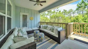 Une véranda couverte avec un ventilateur de plafond, un canapé avec des coussins, un tapis à rayures et une vue sur les arbres.