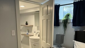Une chambre à coucher comprenant un lit, une table de chevet, une commode, une fenêtre avec des rideaux, un petit bureau avec une lampe, et une salle de bain avec un lavabo et un miroir.