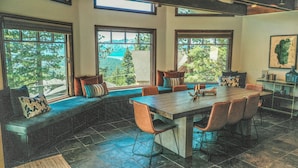Un comedor con una gran mesa de madera, varias sillas y un banco con cojines. Amplias ventanas ofrecen vistas a árboles y montañas.