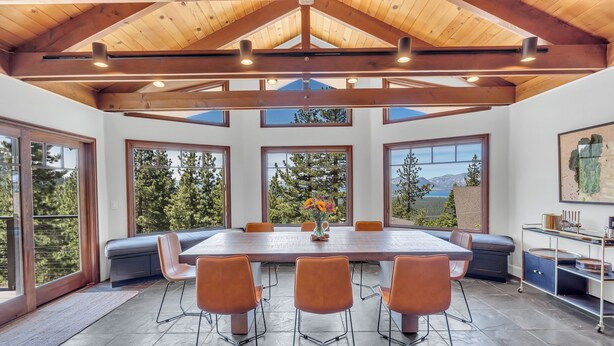 Un comedor amplio con una gran mesa de madera y sillas anaranjadas, rodeado de ventanas que ofrecen vistas a árboles y montañas.