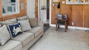 Un salon chaleureux avec un canapé beige, des coussins décoratifs, une petite table d’appoint en bois et une étagère murale ornée d’objets sur le thème de la plage.
